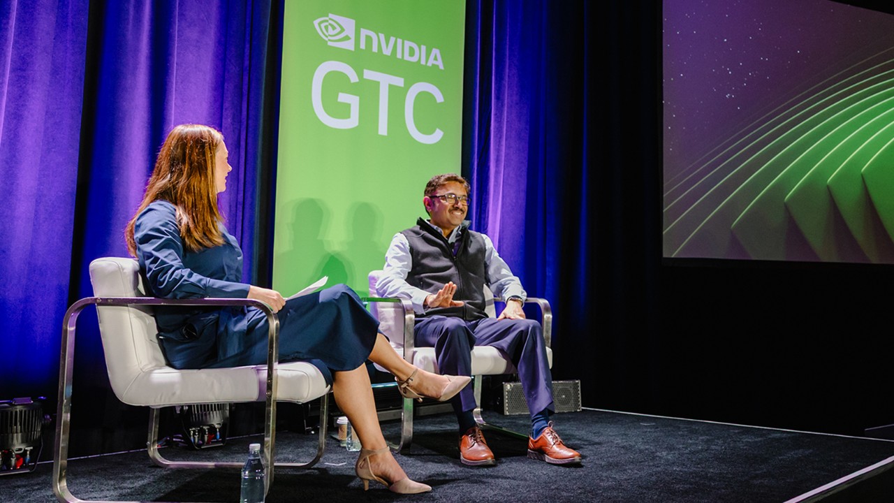 Two speakers sit onstage at an NVIDIA GTC event, talking in front of a green GTC backdrop.