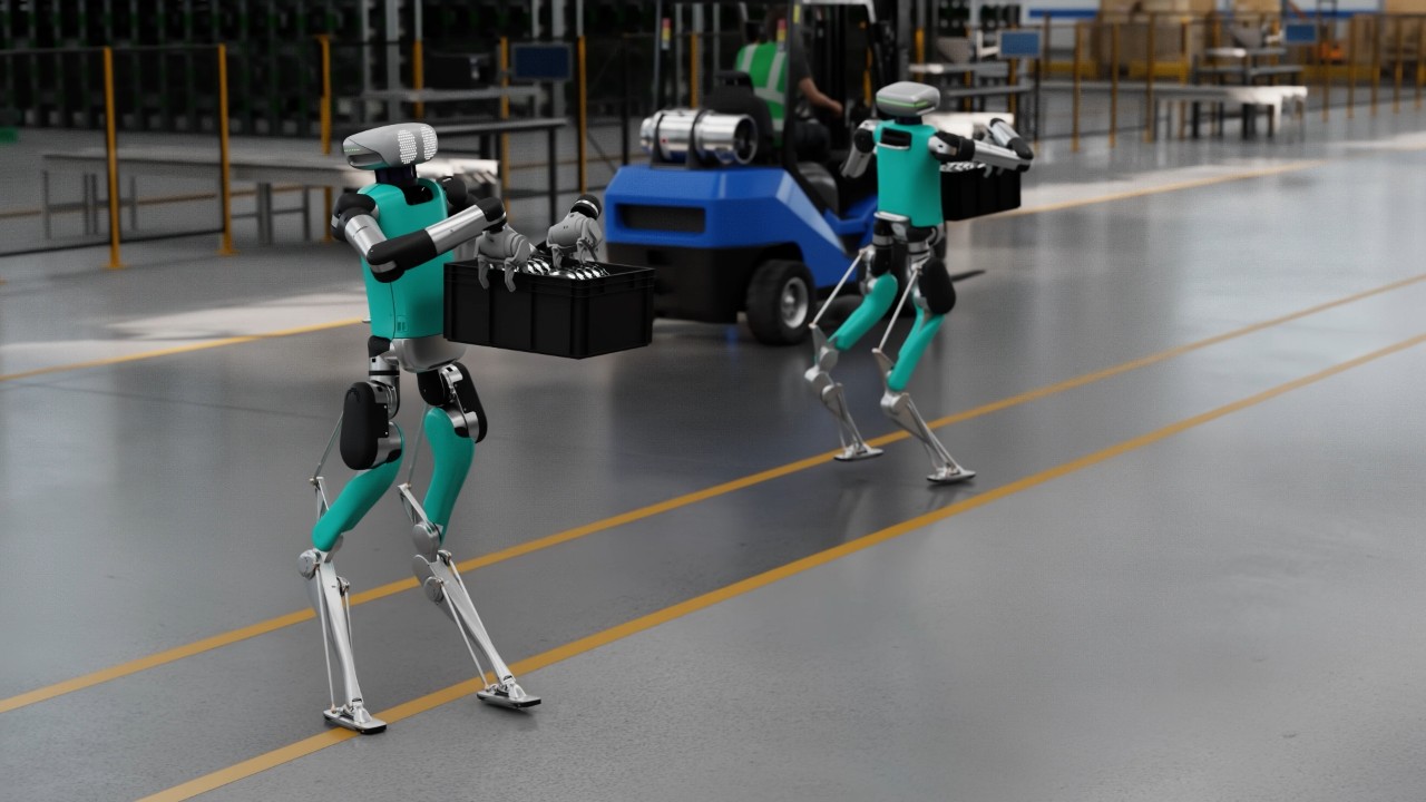 Two teal and black bipedal robots, resembling humans, walking across a factory floor